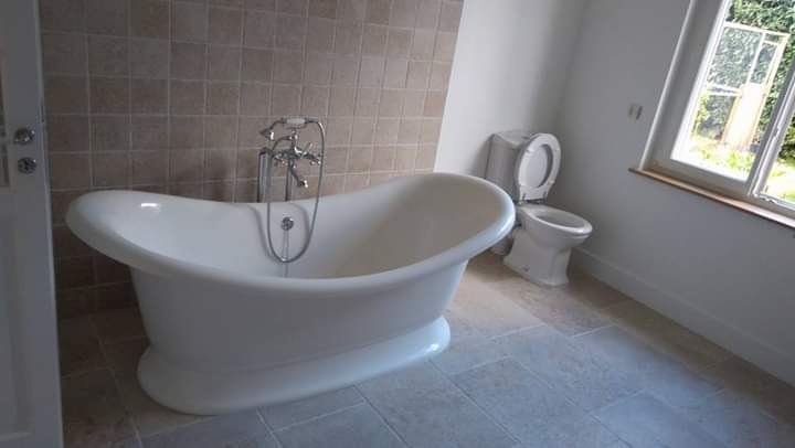 Klasyczna biała wanna wolnostojąca z chromowaną armaturą na tle beżowej ściany z płytek i toalety w jasnym, odnowionym wnętrzu łazienki z oknem.