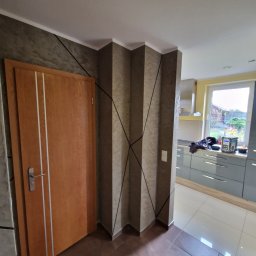 FIRMA HANDLOWO - USŁUGOWA Marcin Grabowski - Wnętrze mieszkania z pomalowanymi ścianami w odcieniach szarości i brązu, z dekoracyjnymi czarnymi liniami. Widoczna drewniana drzwi z białymi wstawkami i fragment kuchni w tle.