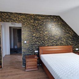 FIRMA HANDLOWO - USŁUGOWA Marcin Grabowski - Sypialnia na poddaszu z dekoracyjną ścianą w kolorze czarno-złotym, widok na sąsiednie pomieszczenie w trakcie remontu, drewniane meble.