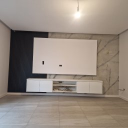 FIRMA HANDLOWO - USŁUGOWA Marcin Grabowski - Nowoczesny salon w trakcie wykańczania: ściana z dekoracyjnymi czarnymi lamelami, płyta imitująca marmur, biała szafka RTV z wiszącym panelem, szare płytki podłogowe. Widoczne wystające kable...