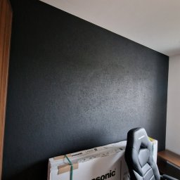 FIRMA HANDLOWO - USŁUGOWA Marcin Grabowski - Czarna ściana z delikatną fakturą, świeżo po malowaniu, z kartonem nowego telewizora Panasonic i krzesłem gamingowym w rogu pokoju.