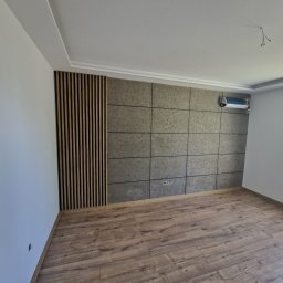 FIRMA HANDLOWO - USŁUGOWA Marcin Grabowski - Pusty pokój po remoncie z panelami ściennymi w odcieniach szarości i drewnianymi lamelami, parkiet w kolorze drewna, grzejnik pod oknem, instalacja klimatyzacji na ścianie, widoczne gniazdka...