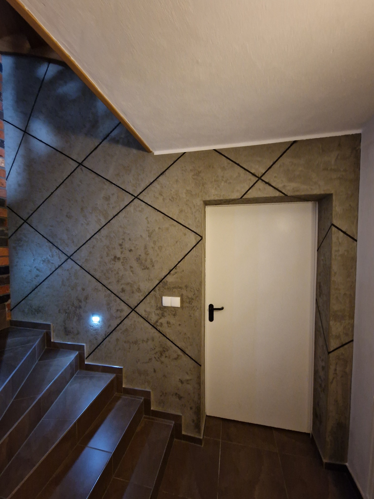 Klatka schodowa z dekoracyjną szarą ścianą w geometryczne wzory, oświetleniem punktowym i białymi drzwiami.