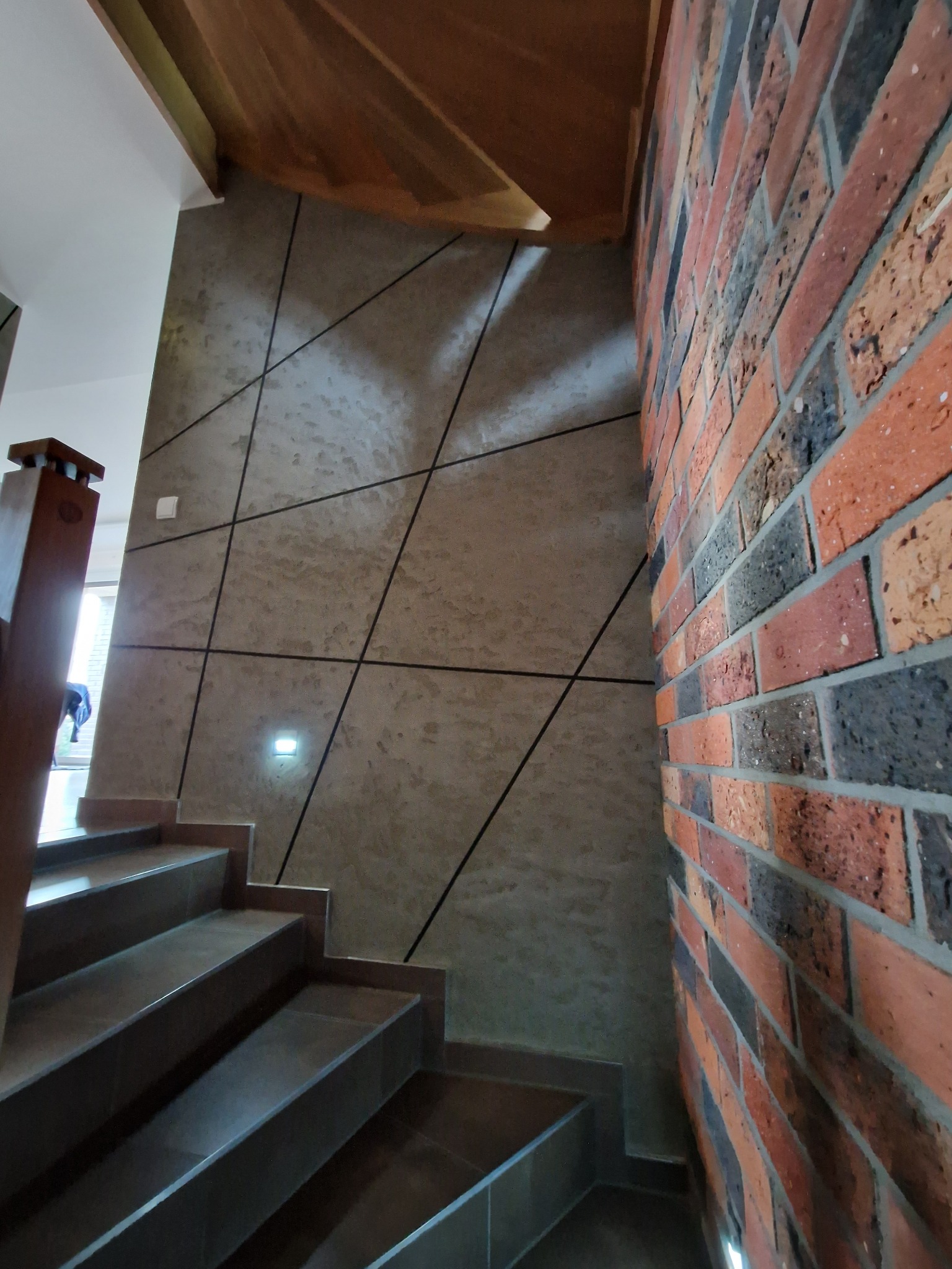 Klatka schodowa z wykończoną ścianą w dekoracyjną strukturę z czarnymi liniami i ścianą z cegły klinkierowej w odcieniach czerwieni i czerni.