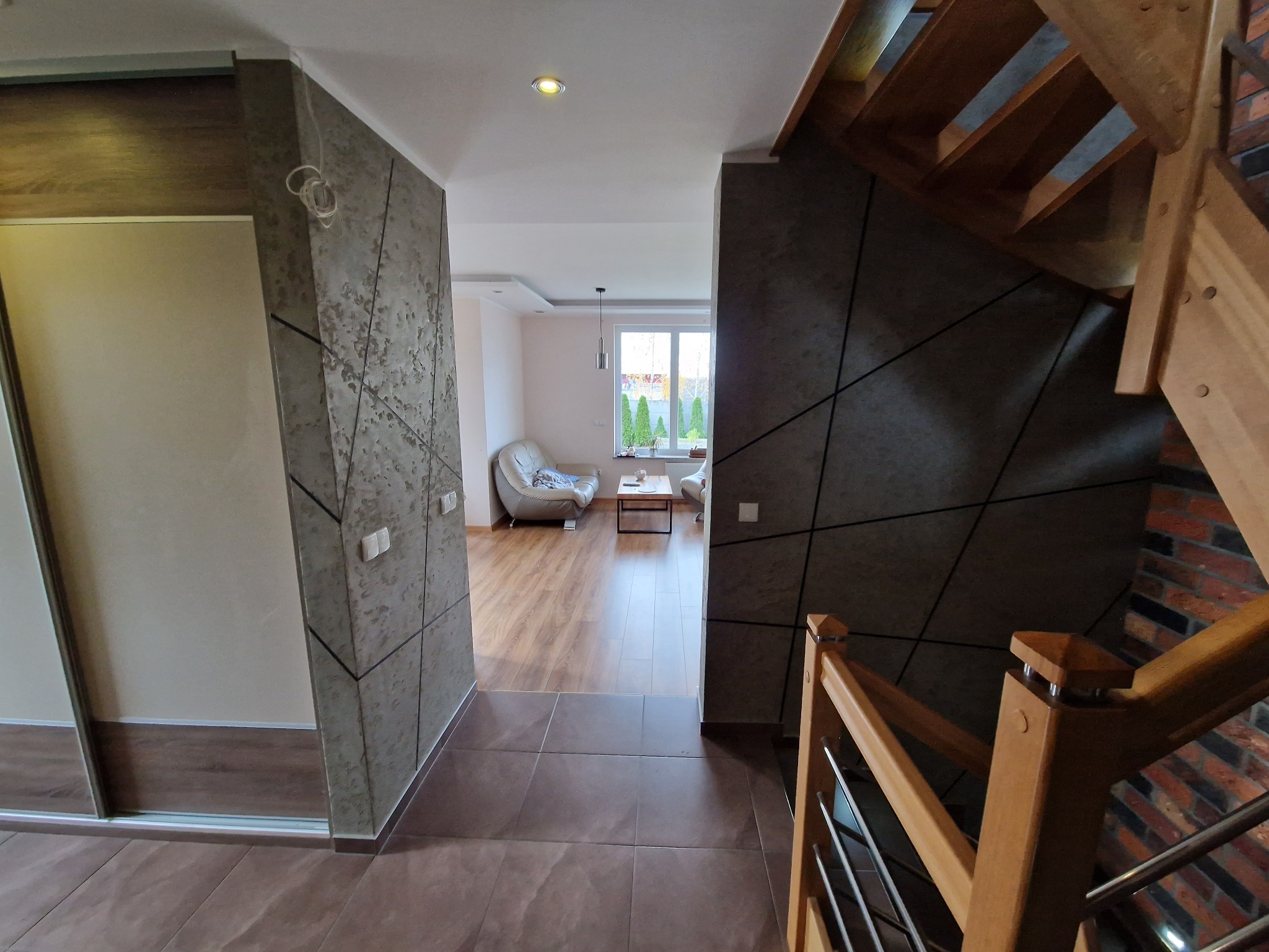 Wnętrze domu z widokiem na salon przez korytarz; na pierwszym planie drewniane schody z metalowymi elementami, po bokach ściany z dekoracyjną strukturą przypominającą beton.