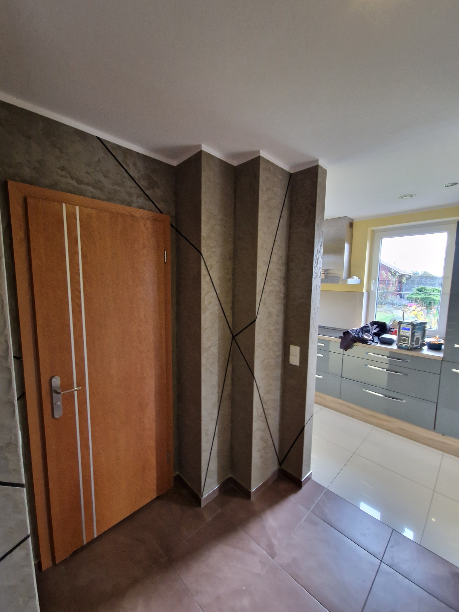 Wnętrze mieszkania z pomalowanymi ścianami w odcieniach szarości i brązu, z dekoracyjnymi czarnymi liniami. Widoczna drewniana drzwi z białymi wstawkami i fragment kuchni w tle.