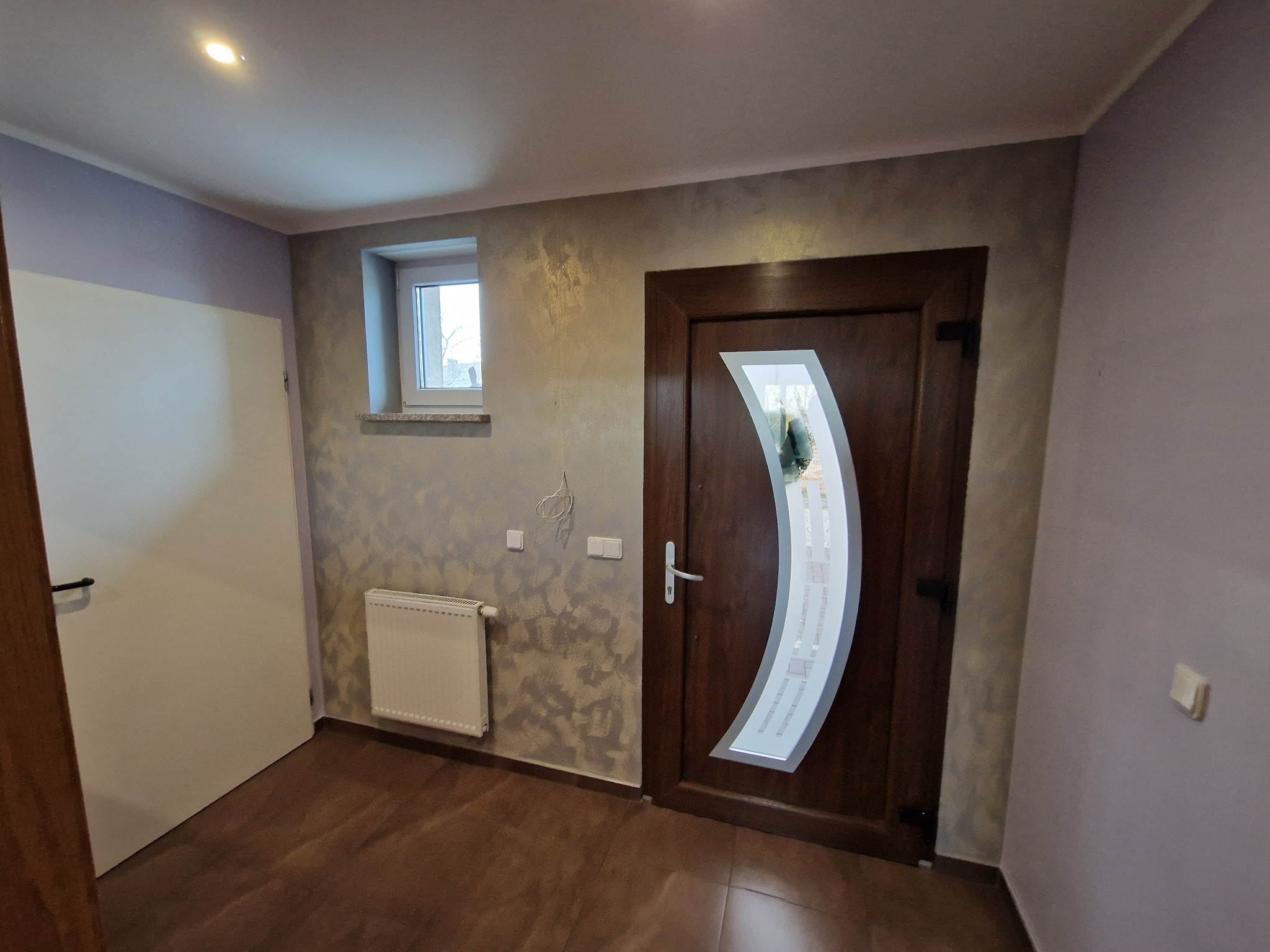 Wnętrze przedpokoju po remoncie: brązowe panele podłogowe, dekoracyjna ściana w odcieniach szarości i złota, biały grzejnik, drewniane drzwi z szybą w kształcie łuku, małe okno z widokiem na zewnątrz.