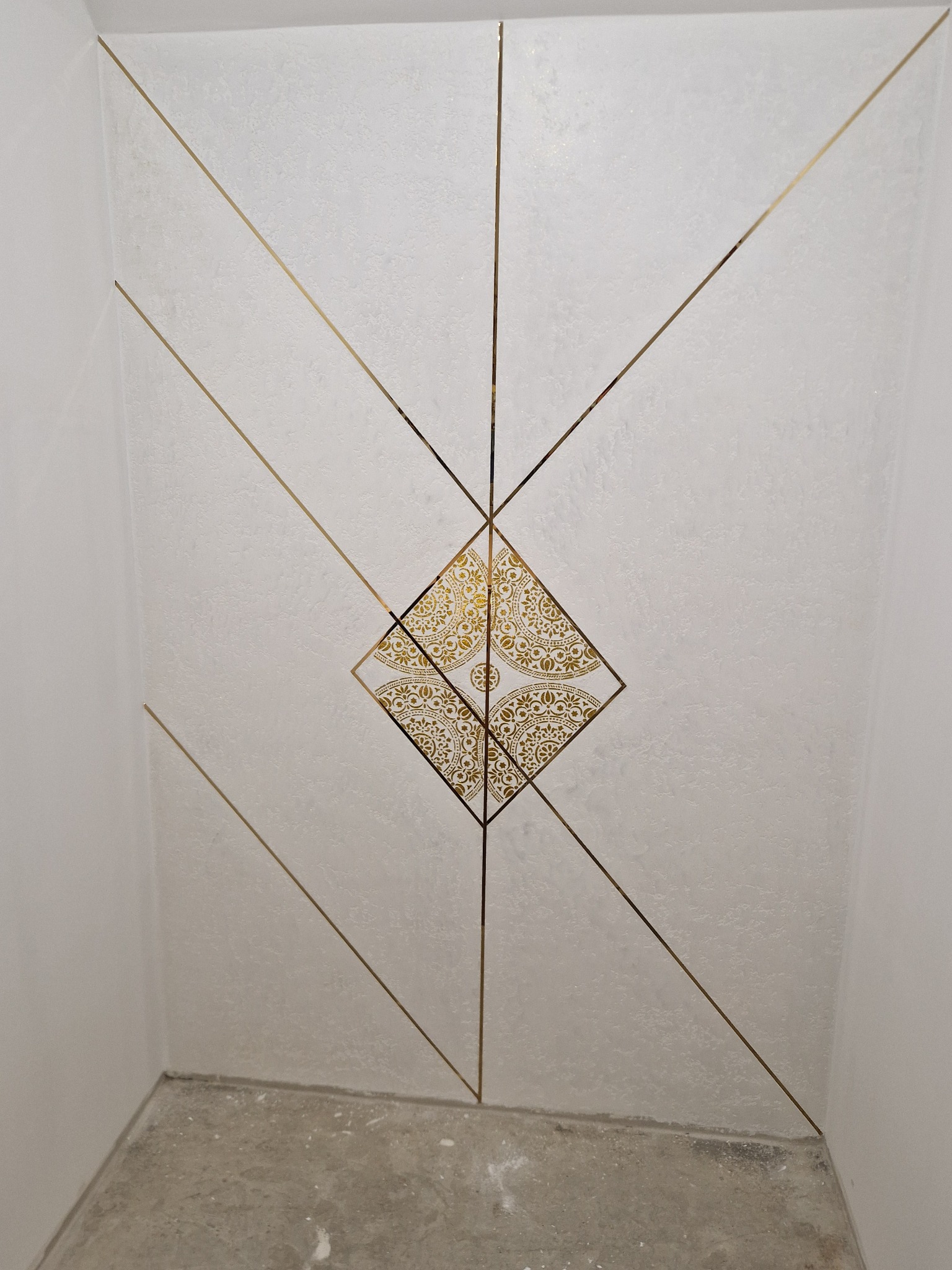 Ściana wykończona białą, strukturalną farbą z dekoracyjnymi, złotymi liniami tworzącymi geometryczny wzór i centralny ornament w kształcie rombu z motywami roślinnymi.
