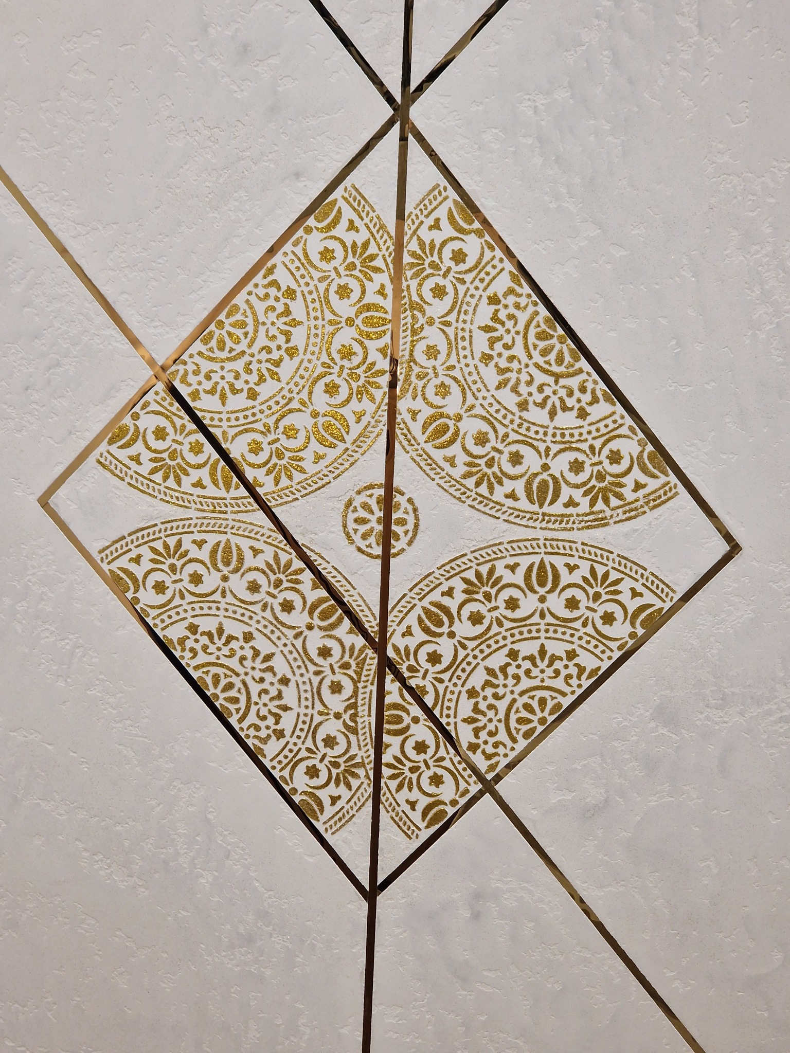 Dekoracyjna ściana z białym tłem o chropowatej fakturze, zdobiona złotym ornamentem w kształcie rombu i cienkimi złotymi liniami krzyżującymi się na powierzchni.
