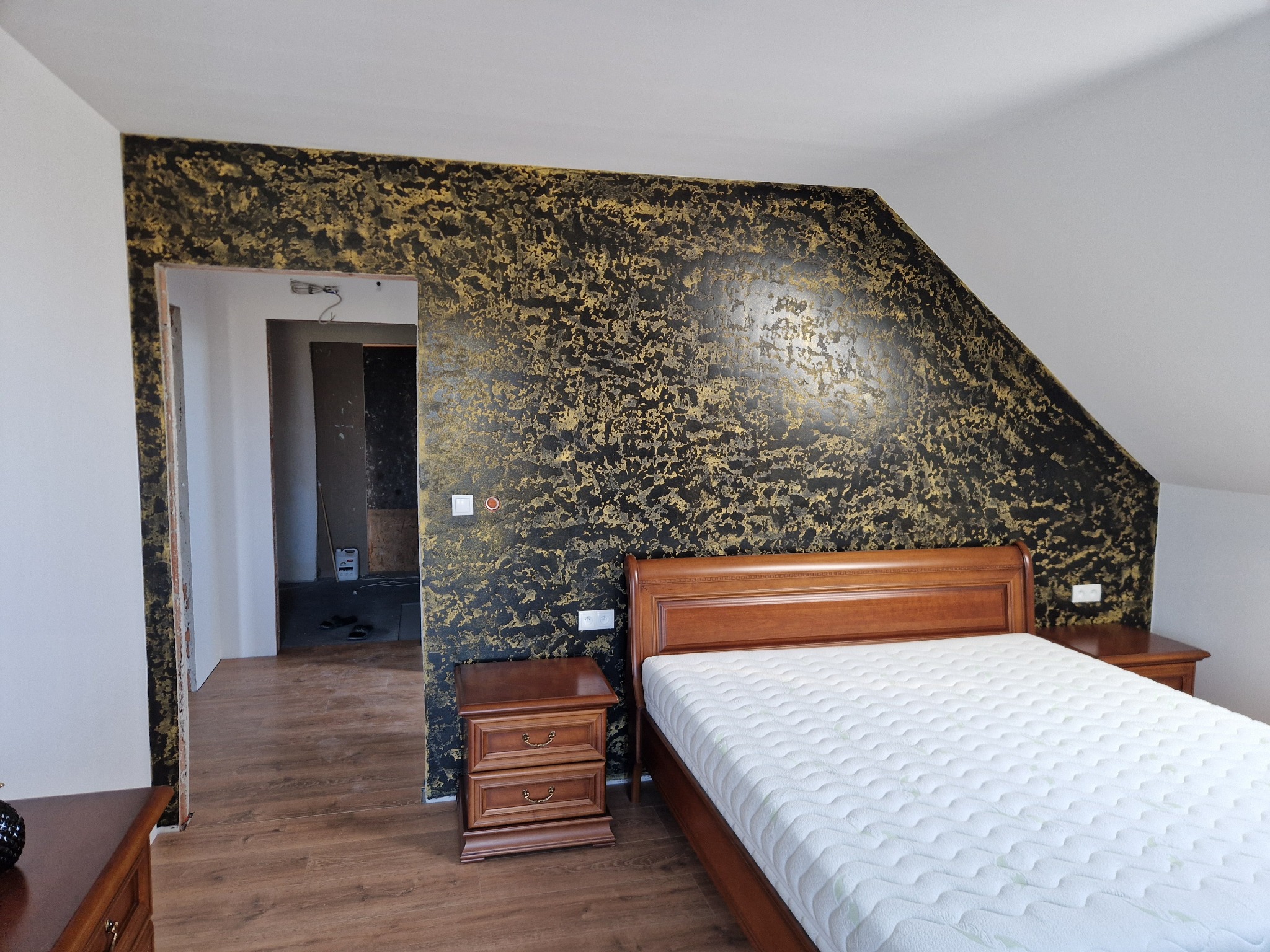 Sypialnia na poddaszu z dekoracyjną ścianą w kolorze czarno-złotym, widok na sąsiednie pomieszczenie w trakcie remontu, drewniane meble.