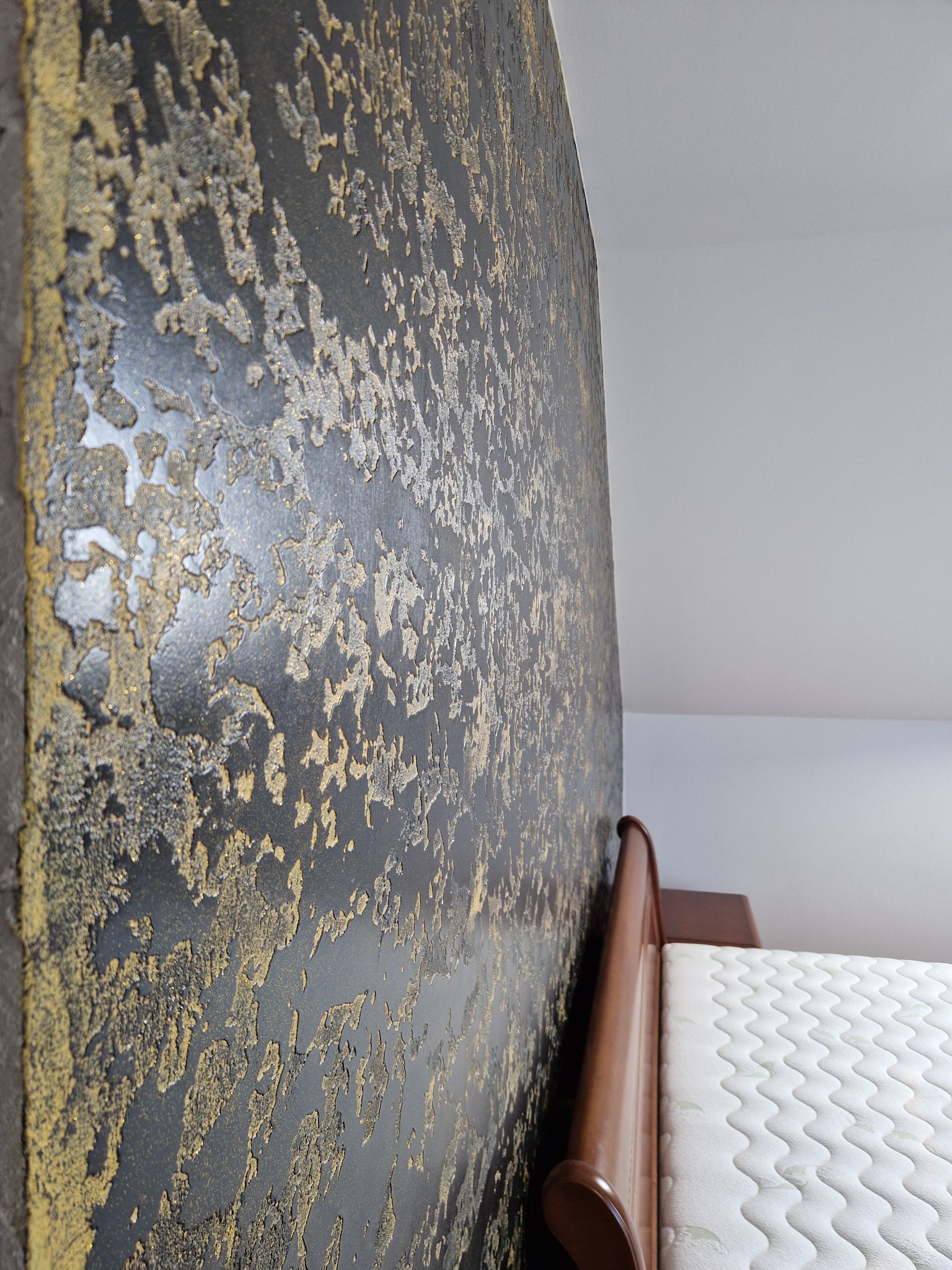 Czarno-złota ściana z dekoracyjną fakturą w sypialni, widoczny fragment drewnianego wezgłowia łóżka i materac