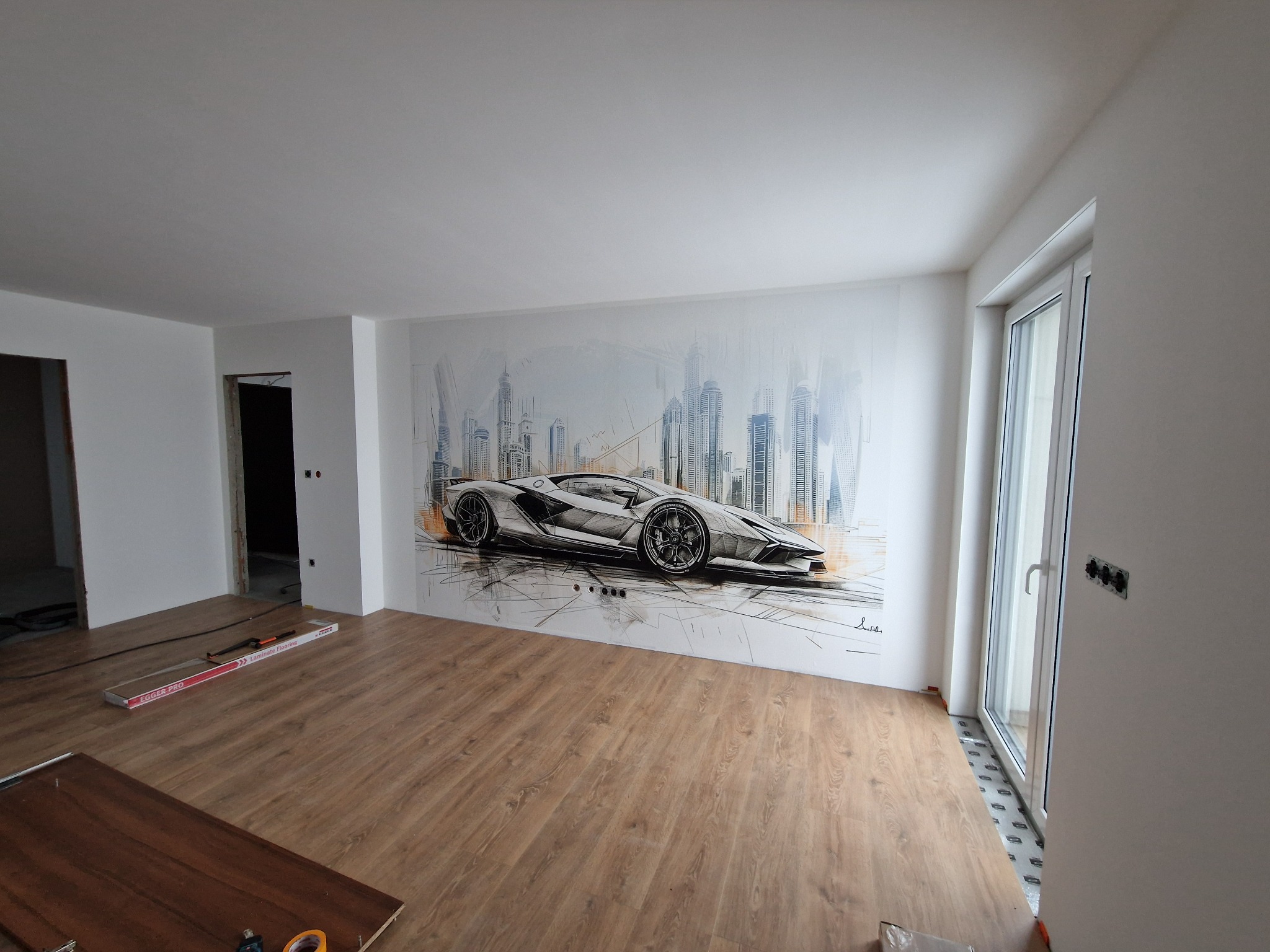 Pokój w trakcie remontu z nową podłogą z paneli i dekoracyjnym malunkiem samochodu sportowego na ścianie.