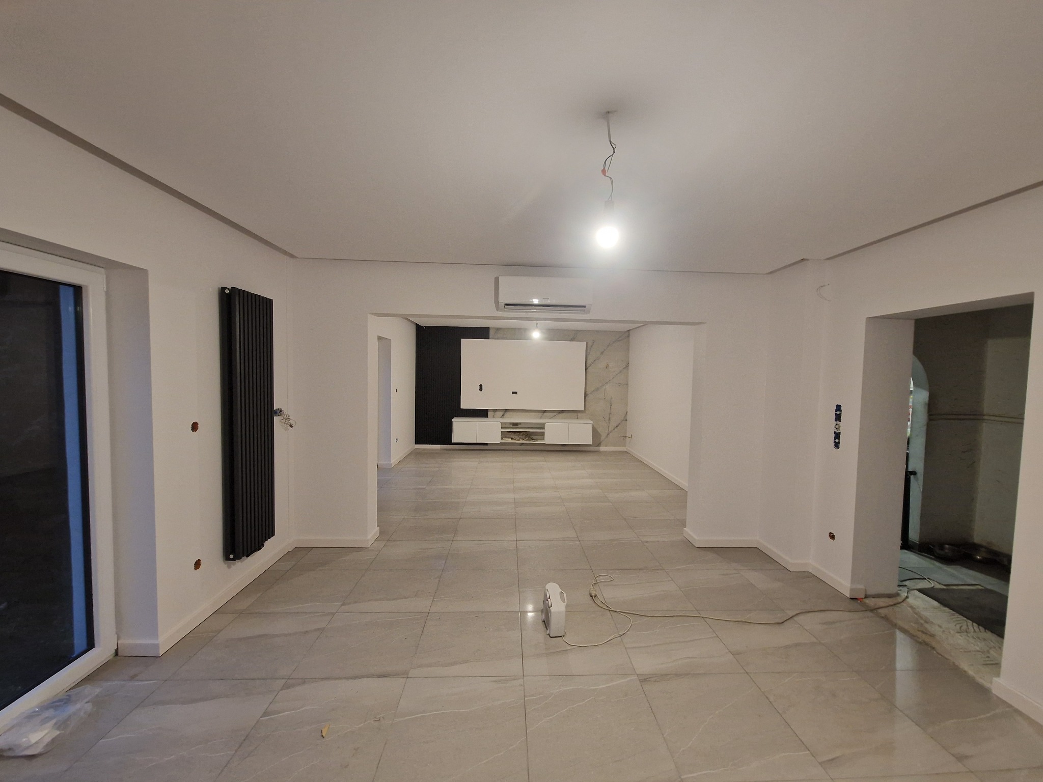 Przestronne wnętrze w trakcie remontu, widoczne płytki podłogowe imitujące marmur, grzejnik panelowy na ścianie, białe ściany z widocznymi puszkami elektrycznymi, oraz system klimatyzacji pod sufitem.