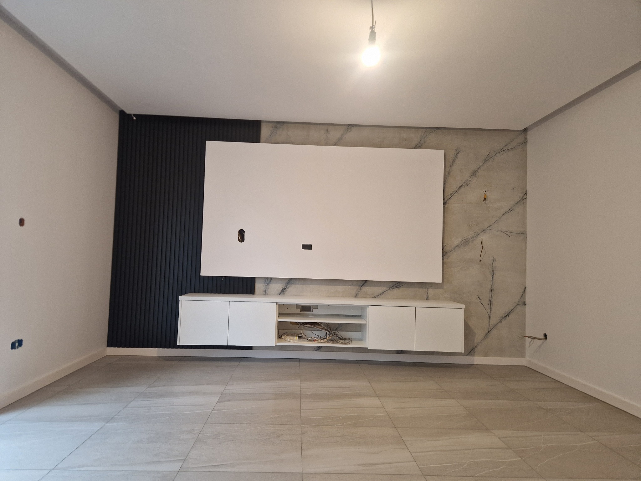 Nowoczesny salon w trakcie wykańczania: ściana z dekoracyjnymi czarnymi lamelami, płyta imitująca marmur, biała szafka RTV z wiszącym panelem, szare płytki podłogowe. Widoczne wystające kable...