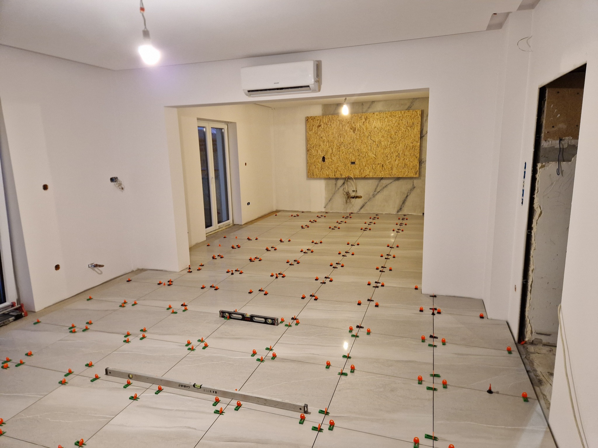 Podłoga w trakcie układania płytek ceramicznych, widoczne poziomice, krzyżyki dystansowe i sznurki wyznaczające linie proste, surowy stan ścian.