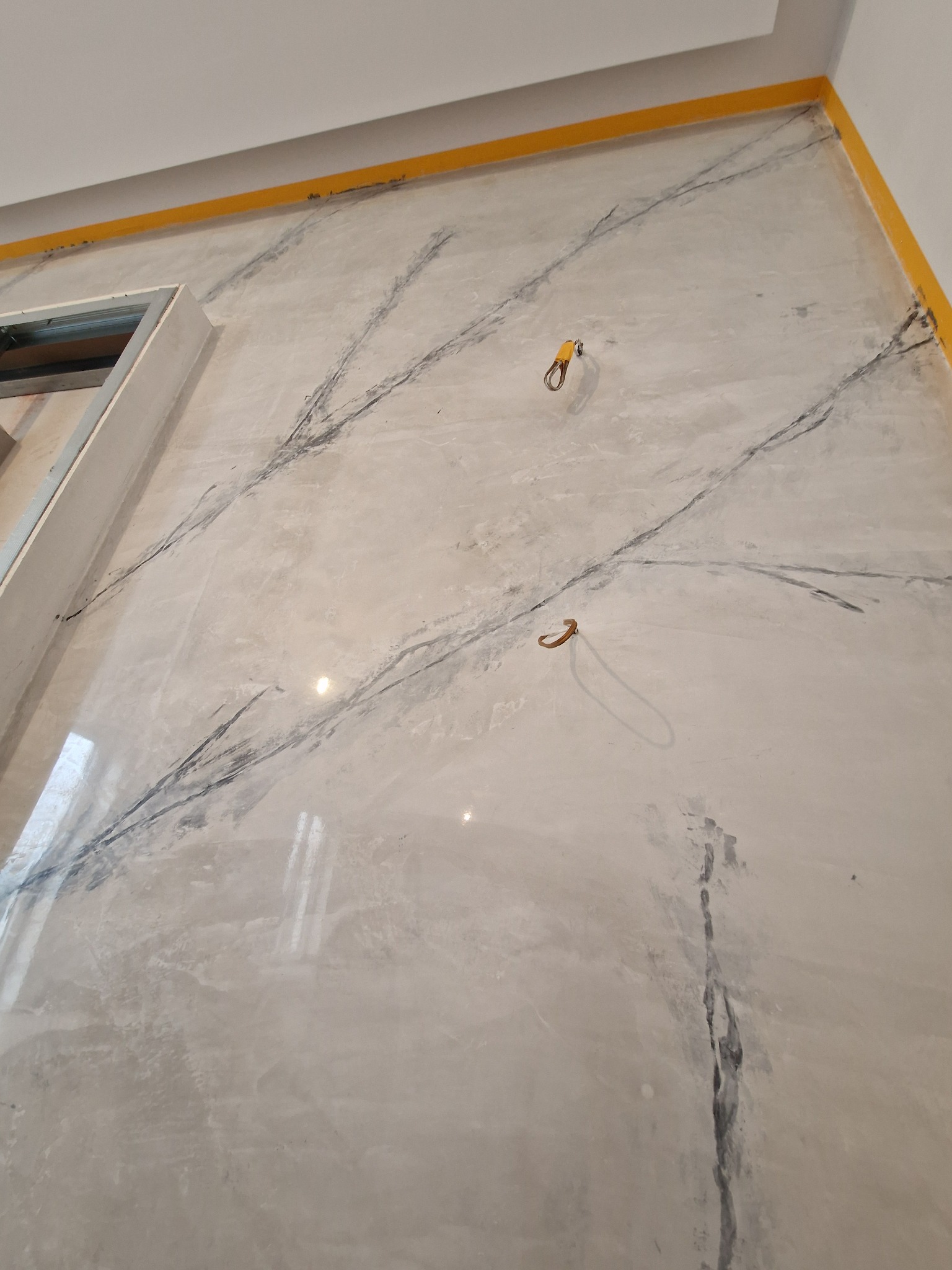 Ściana wewnętrzna z dekoracyjną, marmurkową fakturą, zabezpieczona żółtą taśmą malarską przy suficie i narożnikach, widoczne dwa uchwyty na ścianie i fragment otworu w ścianie.