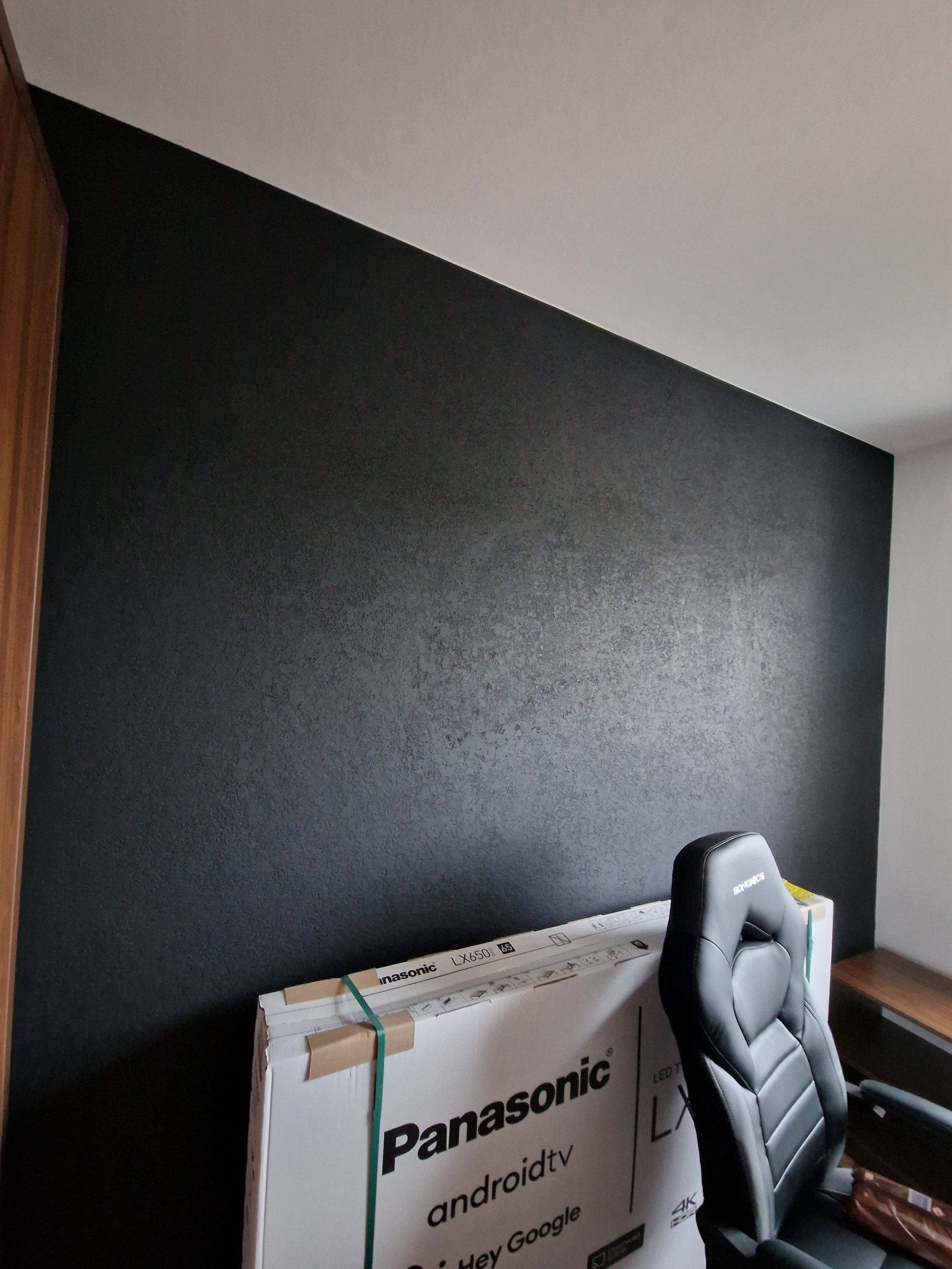 Czarna ściana z delikatną fakturą, świeżo po malowaniu, z kartonem nowego telewizora Panasonic i krzesłem gamingowym w rogu pokoju.