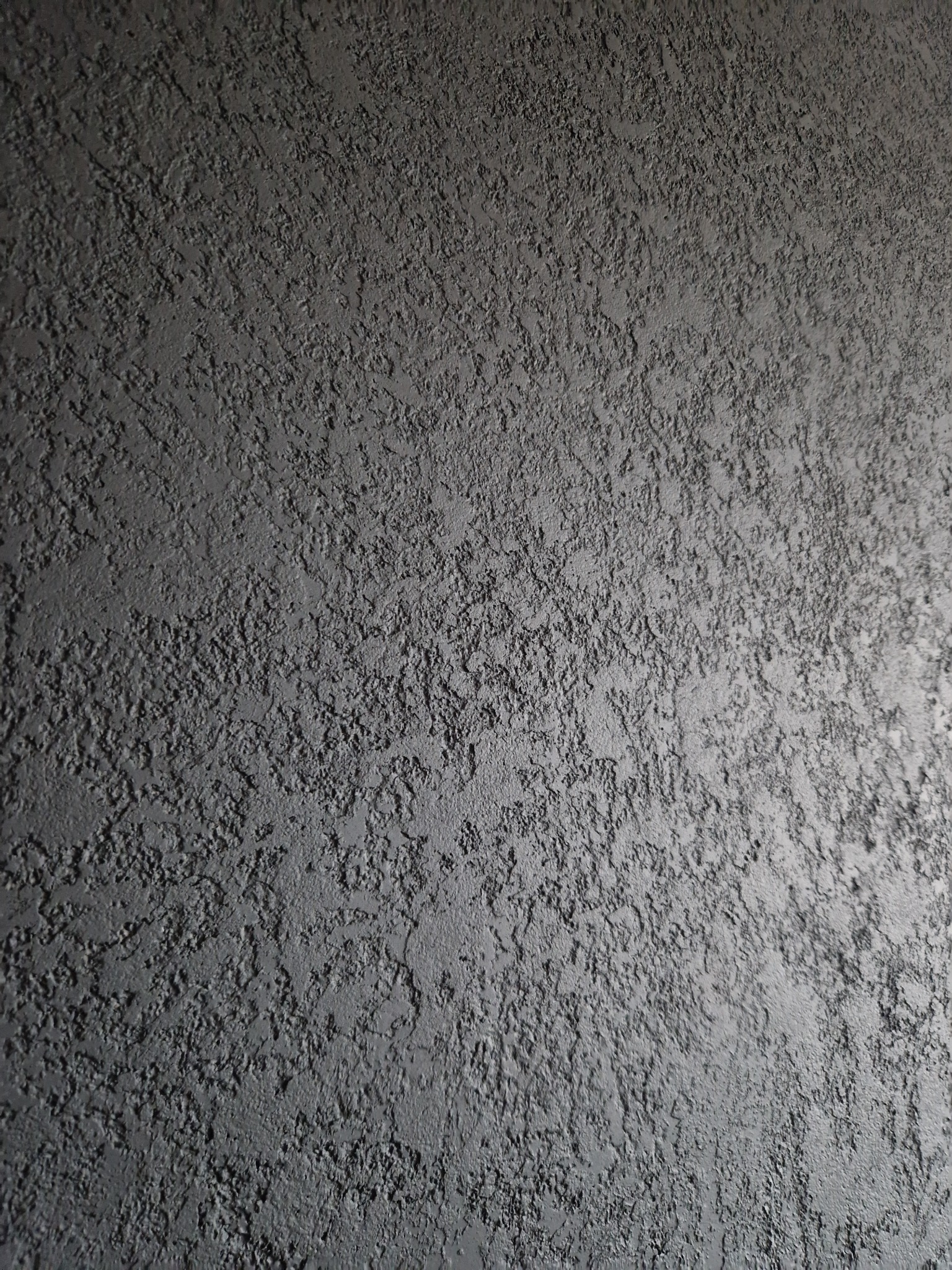 Szara ściana pokryta grubą, strukturalną farbą, tworzącą nierówną, chropowatą powierzchnię z widocznymi zagłębieniami i wypukłościami.