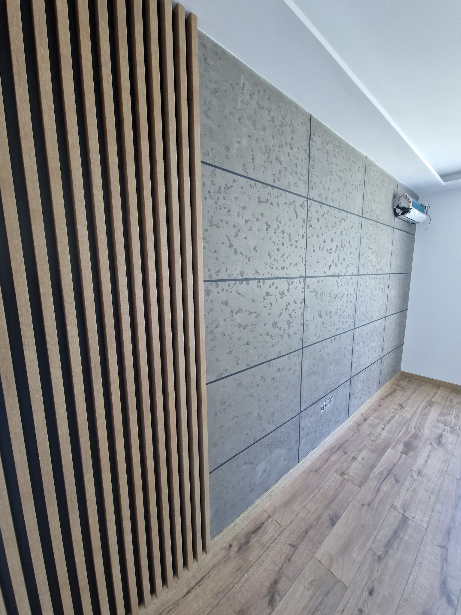 Nowoczesne wnętrze z panelami ściennymi imitującymi beton, drewnianymi lamelami dekoracyjnymi, jasną podłogą i klimatyzatorem na ścianie.