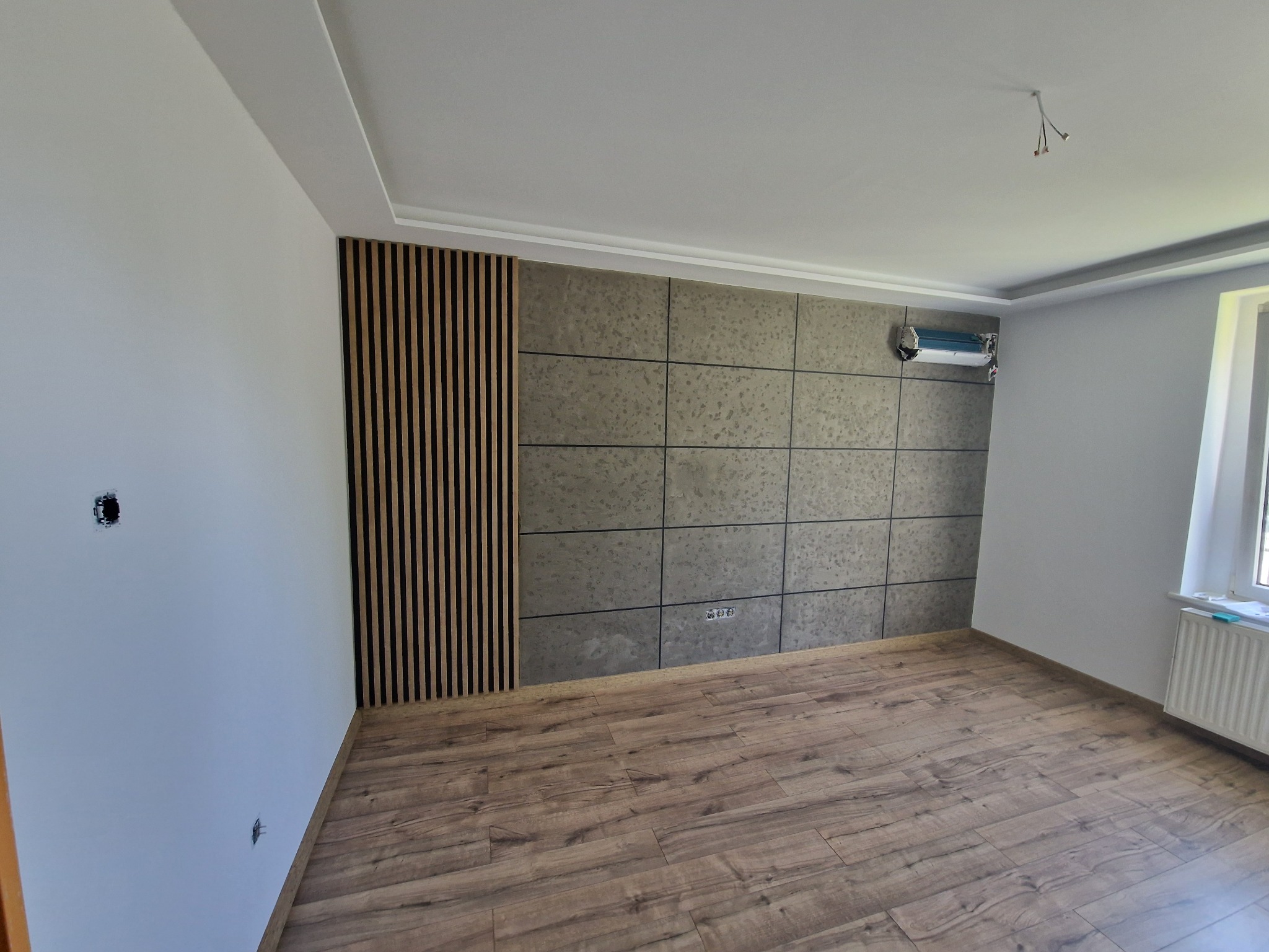 Pusty pokój po remoncie z panelami ściennymi w odcieniach szarości i drewnianymi lamelami, parkiet w kolorze drewna, grzejnik pod oknem, instalacja klimatyzacji na ścianie, widoczne gniazdka...