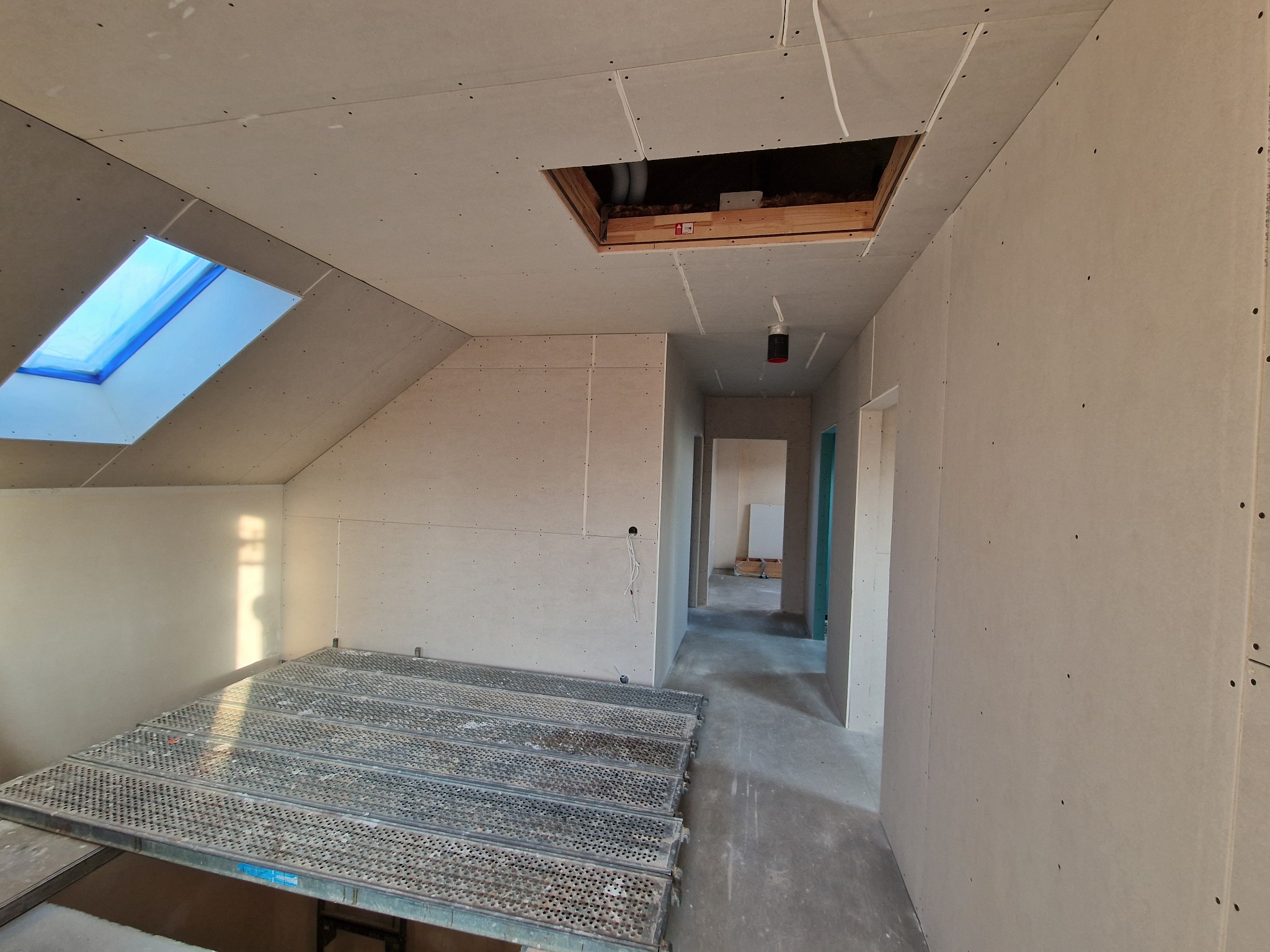 Poddasze w trakcie wykańczania płytami kartonowo-gipsowymi, widoczne rusztowanie, okno dachowe zabezpieczone folią oraz otwór w suficie z instalacją.