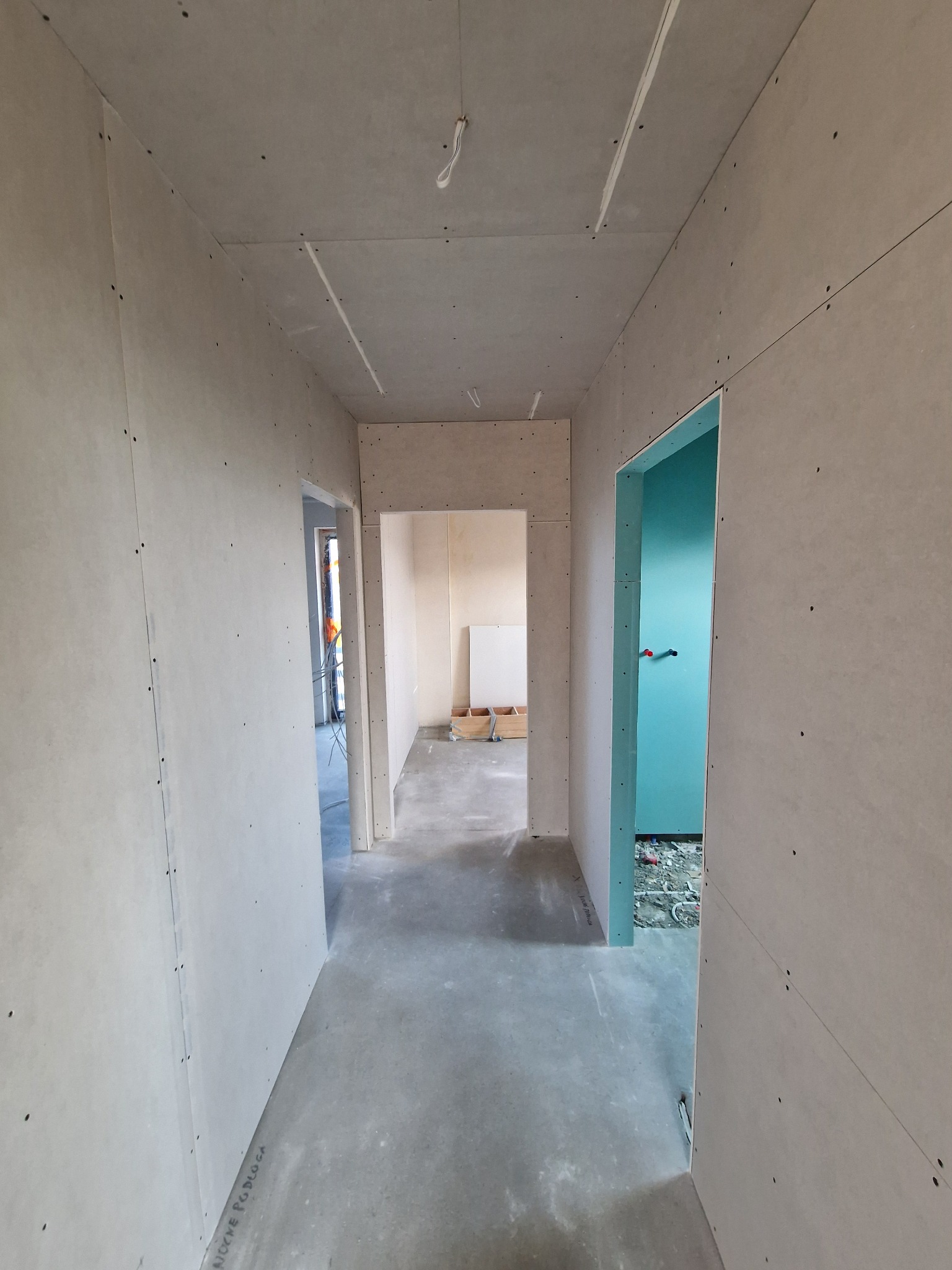 Korytarz w trakcie remontu z widocznymi płytami kartonowo-gipsowymi na ścianach i suficie, niezagruntowane, widoczne łączenia i śruby, w głębi widoczne otwarte drzwi do wyremontowanej łazienki...
