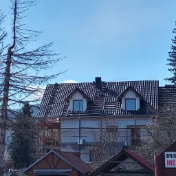 Dom z częściowo zdjętym starym pokryciem dachowym, widoczne łaty, rusztowania i okna dachowe typu lukarna.