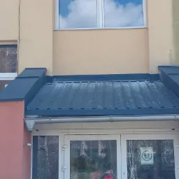 Zadaszenie nad wejściem do budynku z blachy w kolorze grafitowym, z widocznymi rynnami i obróbkami blacharskimi, na tle beżowej elewacji budynku.