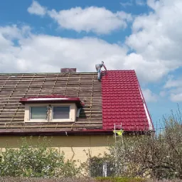 Montaż czerwonej blachodachówki na dachu domu jednorodzinnego, widoczna drabina oparta o dach i dekarz w trakcie pracy na tle błękitnego nieba z chmurami.