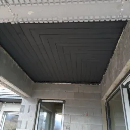 Poddasze w trakcie ocieplania, widoczne szare płyty styropianowe z charakterystycznym wzorem i czarna izolacja termiczna ułożona w geometryczny wzór, fragment okna.