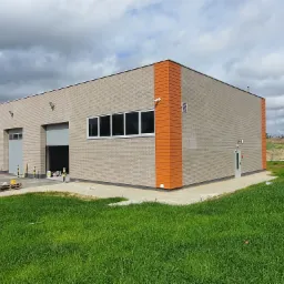 Nowoczesny budynek przemysłowy z elewacją z cegły i pomarańczowymi akcentami, widoczne otwarte bramy garażowe, trawnik na pierwszym planie i pochmurne niebo w tle.