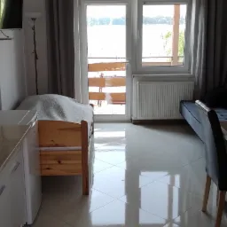 Jasne wnętrze pokoju z widokiem na jezioro przez balkon; błyszcząca podłoga odbija światło, w tle pojedyncze ł&oacute;żko i sofa.