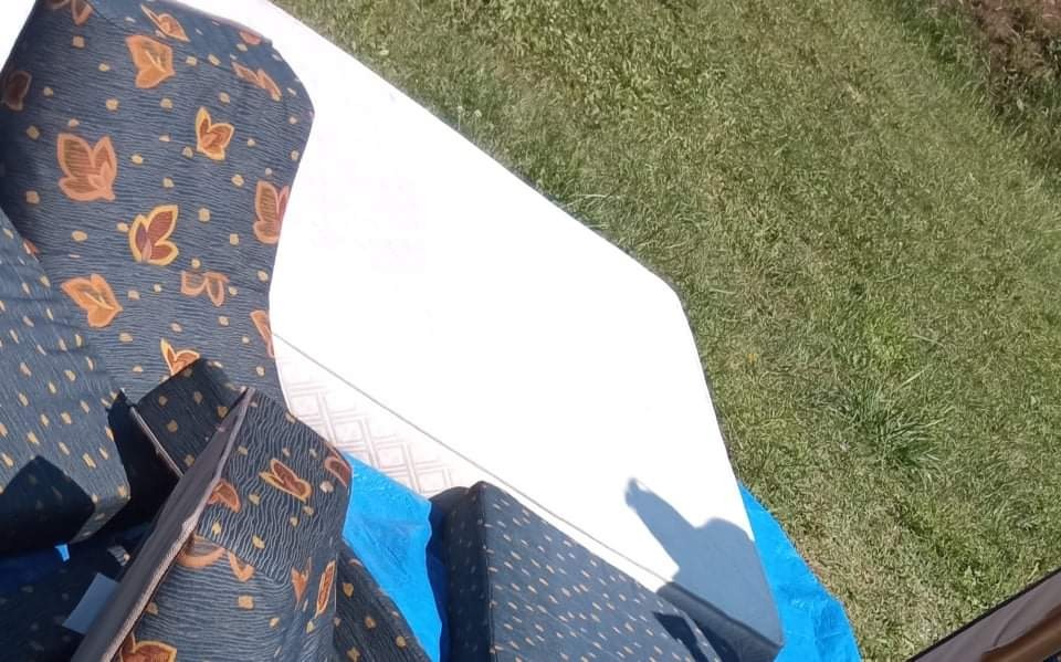 Wyjęte z przyczepy kempingowej poduszki z tapicerką w niebiesko-brązowe wzory oraz biały materac leżą na niebieskiej folii na trawie.