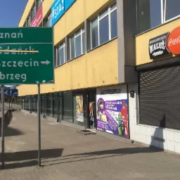 Fasada budynku z szyldem 'Wypożyczalnia Strojów i Przebrań' w Koszalinie i Słupsku, widoczna z chodnika z kostki brukowej, obok drogowskaz na Poznań, Gdańsk, Szczecin i Kołobrzeg.