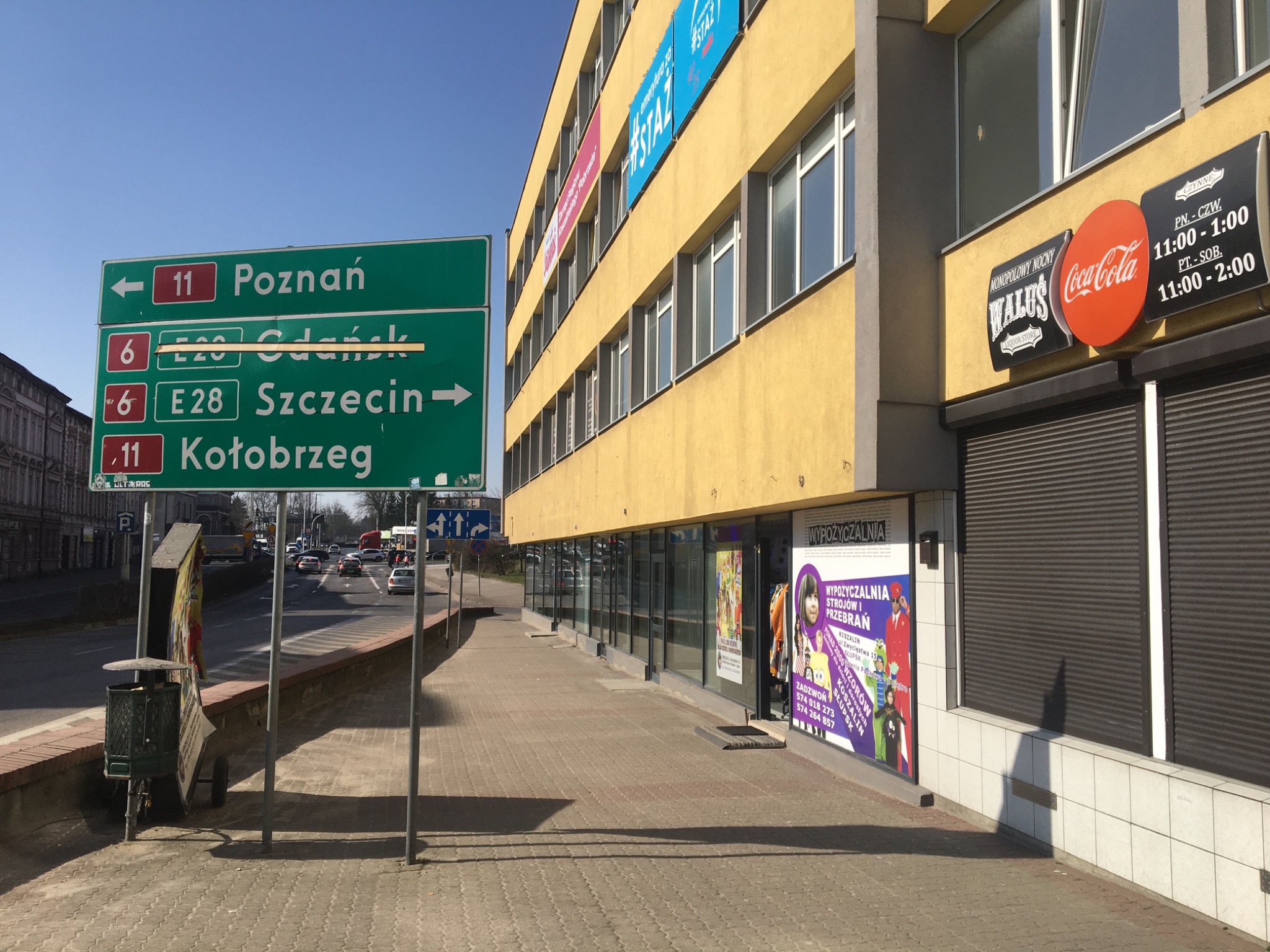 Żółty budynek z plakatem reklamującym wypożyczalnię kostiumów i przebrań, obok chodnik i znak drogowy wskazujący kierunki na Poznań, Gdańsk, Szczecin i Kołobrzeg.