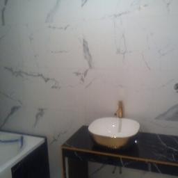 Artur Berdnik - Umywalka w kolorze białym i złotym, osadzona na czarnym blacie z marmurowym wzorem, na tle ściany wyłożonej białymi płytkami imitującymi marmur, widoczna bateria w kolorze złotym i fragment wanny...