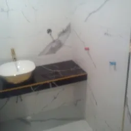 Umywalka nablatowa w kolorze białym ze złotym wykończeniem, zamontowana na czarnym blacie z marmurkowym wzorem, na tle białych płytek z szarymi żyłami w łazience.