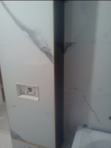 Biała ściana wyłożona płytkami imitującymi marmur z wystającym gniazdkiem elektrycznym, widoczne niedoskonałości i zabrudzenia na powierzchni płytek.