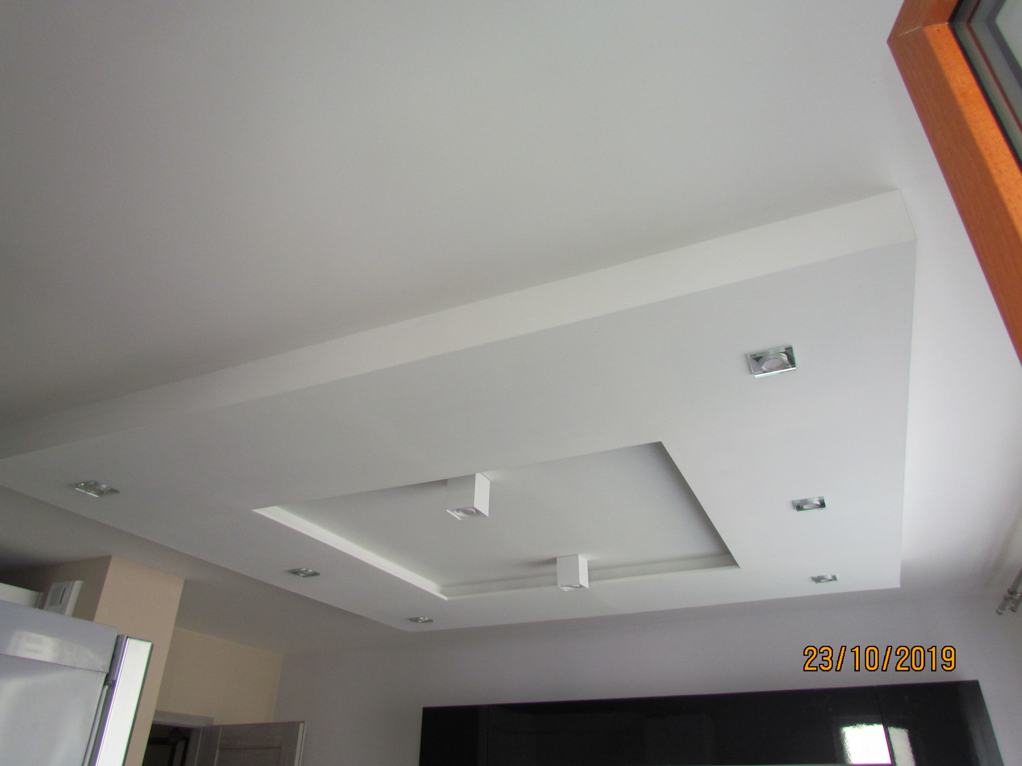 Podwieszany sufit w kształcie kwadratu z wbudowanym oświetleniem punktowym, widok z dołu, fragment okna w prawym górnym rogu, data 23/10/2019 w prawym dolnym rogu.