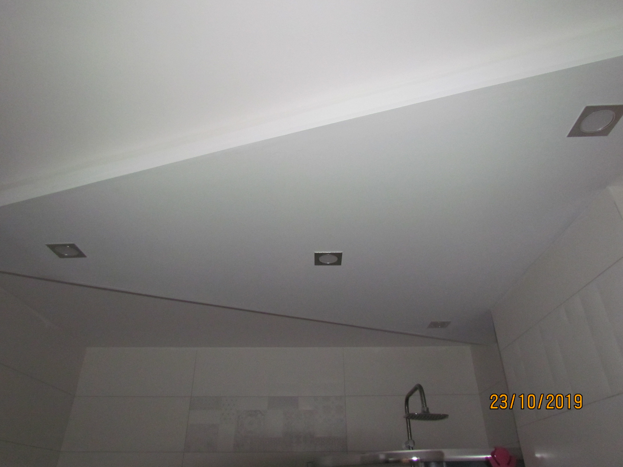 Wykończony sufit w łazience z trzema wbudowanymi, kwadratowymi lampami halogenowymi, widoczne płytki ścienne i bateria prysznicowa.