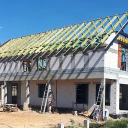 Budowa domu jednorodzinnego z widoczną konstrukcją drewnianą dachu, ściany z bloczków, drabiny oparta o ściany, paleta na ziemi, błękitne niebo w tle.