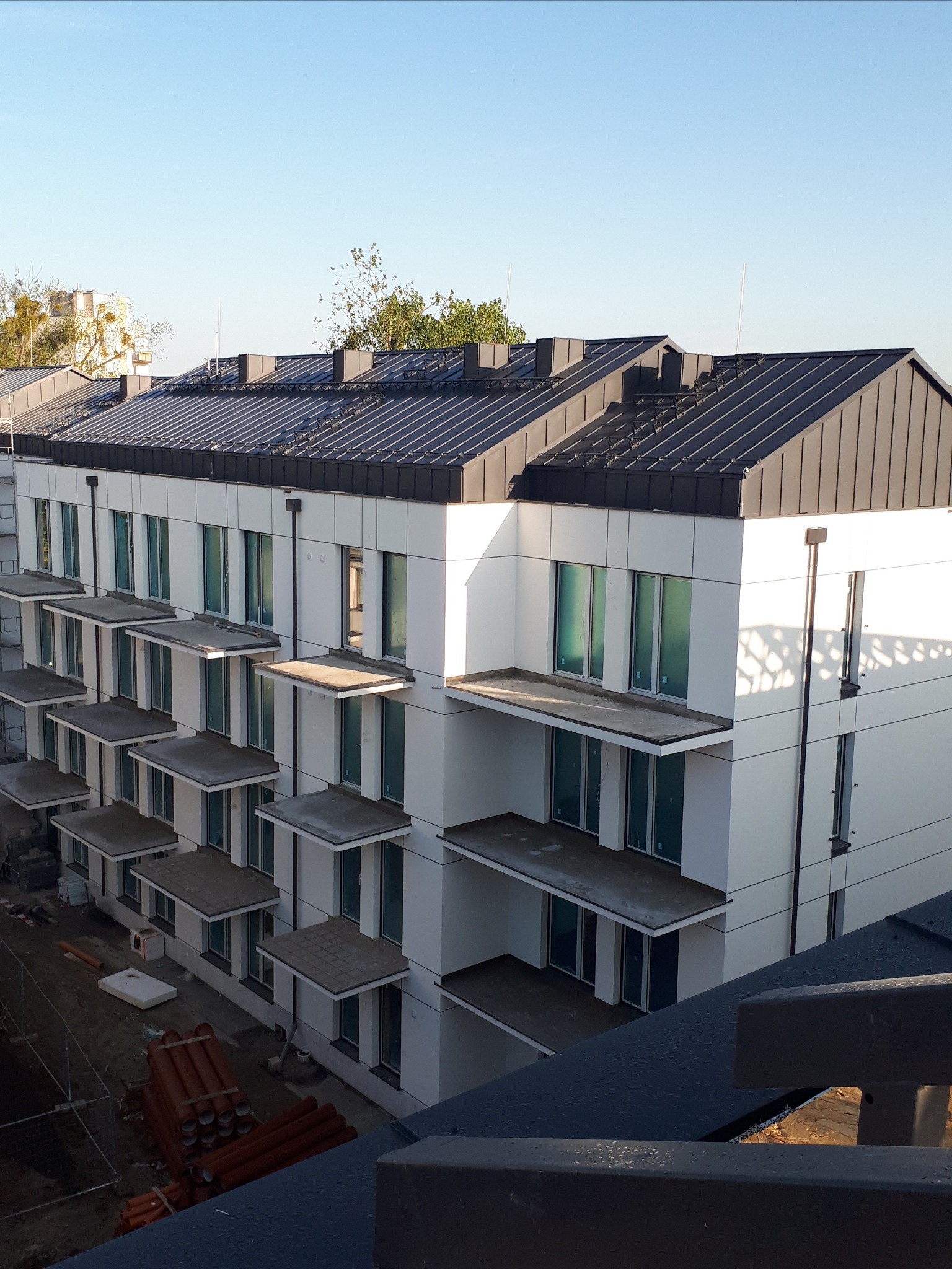 Nowoczesny budynek wielorodzinny z grafitowym dachem z blachy na rąbek stojący, widok z góry na ukończony dach i elewację, w tle błękitne niebo.