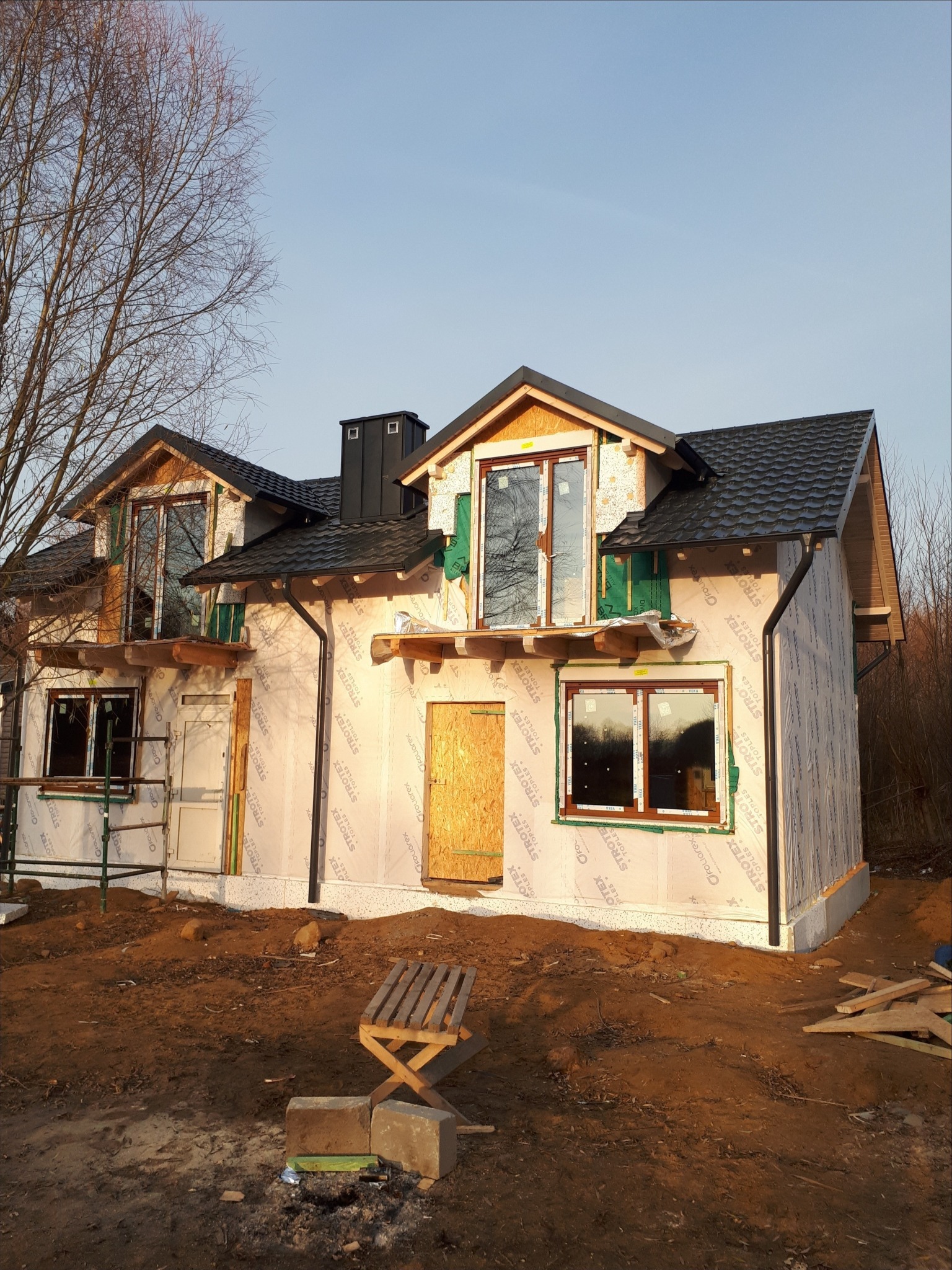 Dom w trakcie budowy, widoczny dach z czarnej blachy falistej, okna, oraz ściany pokryte folią izolacyjną. Na pierwszym planie prowizoryczne siedzisko z palet i cegieł.