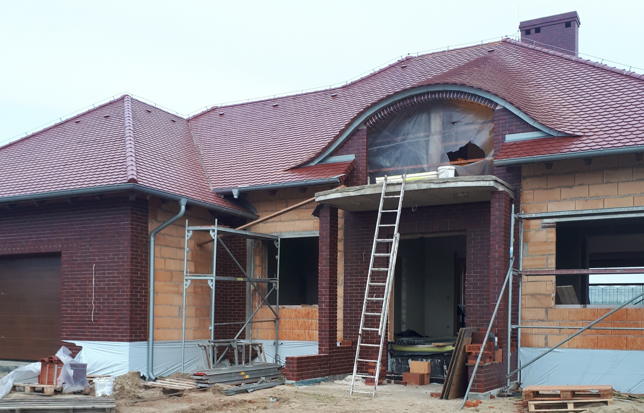 Dom jednorodzinny w trakcie budowy, widoczny nowy dach z czerwonej dachówki, ściany z cegły klinkierowej, rusztowania i drabina oparta o balkon, okno zabezpieczone folią.