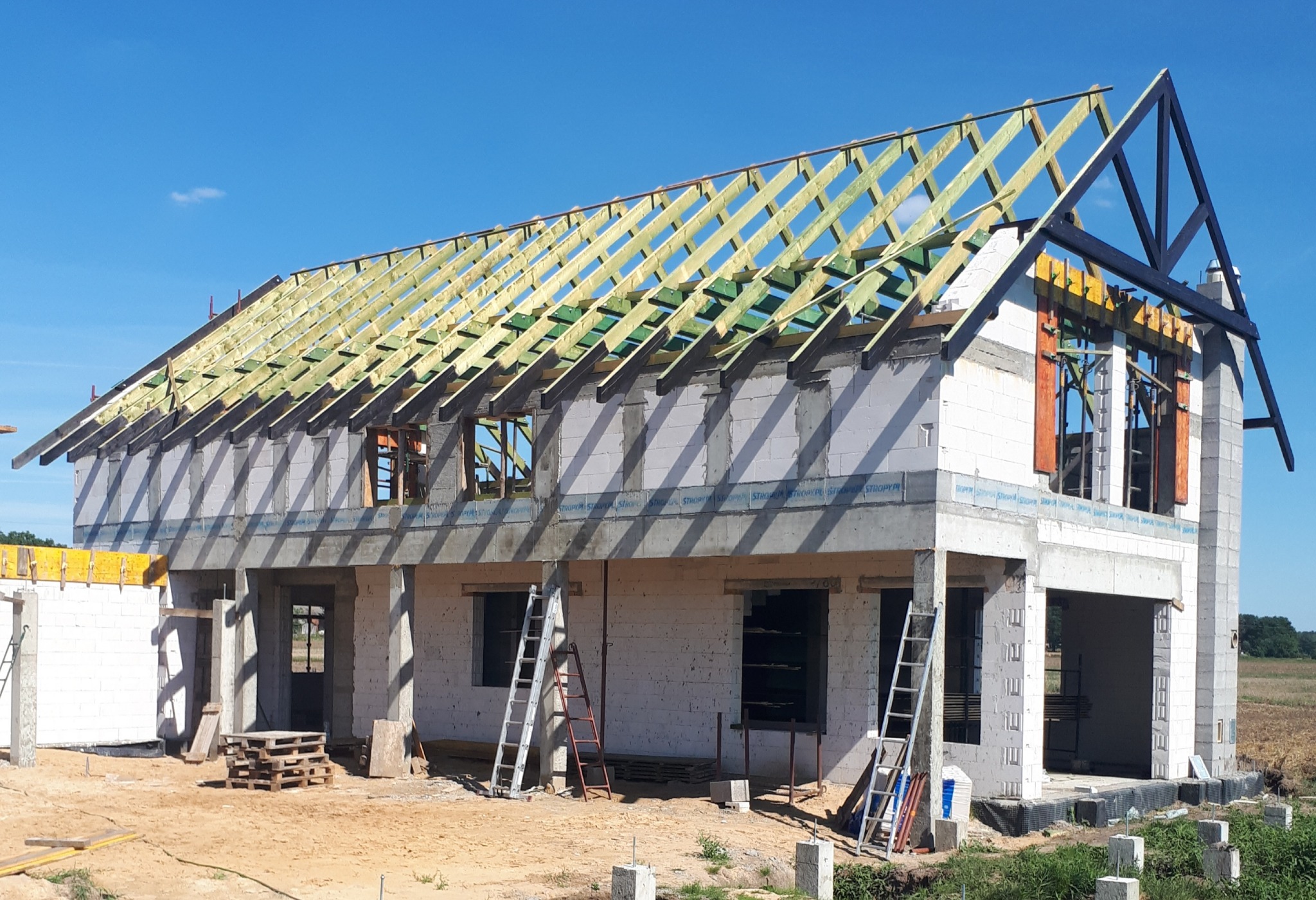 Budowa domu jednorodzinnego z widoczną konstrukcją drewnianą dachu, ściany z bloczków, drabiny oparta o ściany, paleta na ziemi, błękitne niebo w tle.