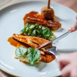 Elegancko podane danie z pieczonego mięsa w sosie, dekorowane zielenią i kwiatami, krojone nożem na jasnym talerzu.