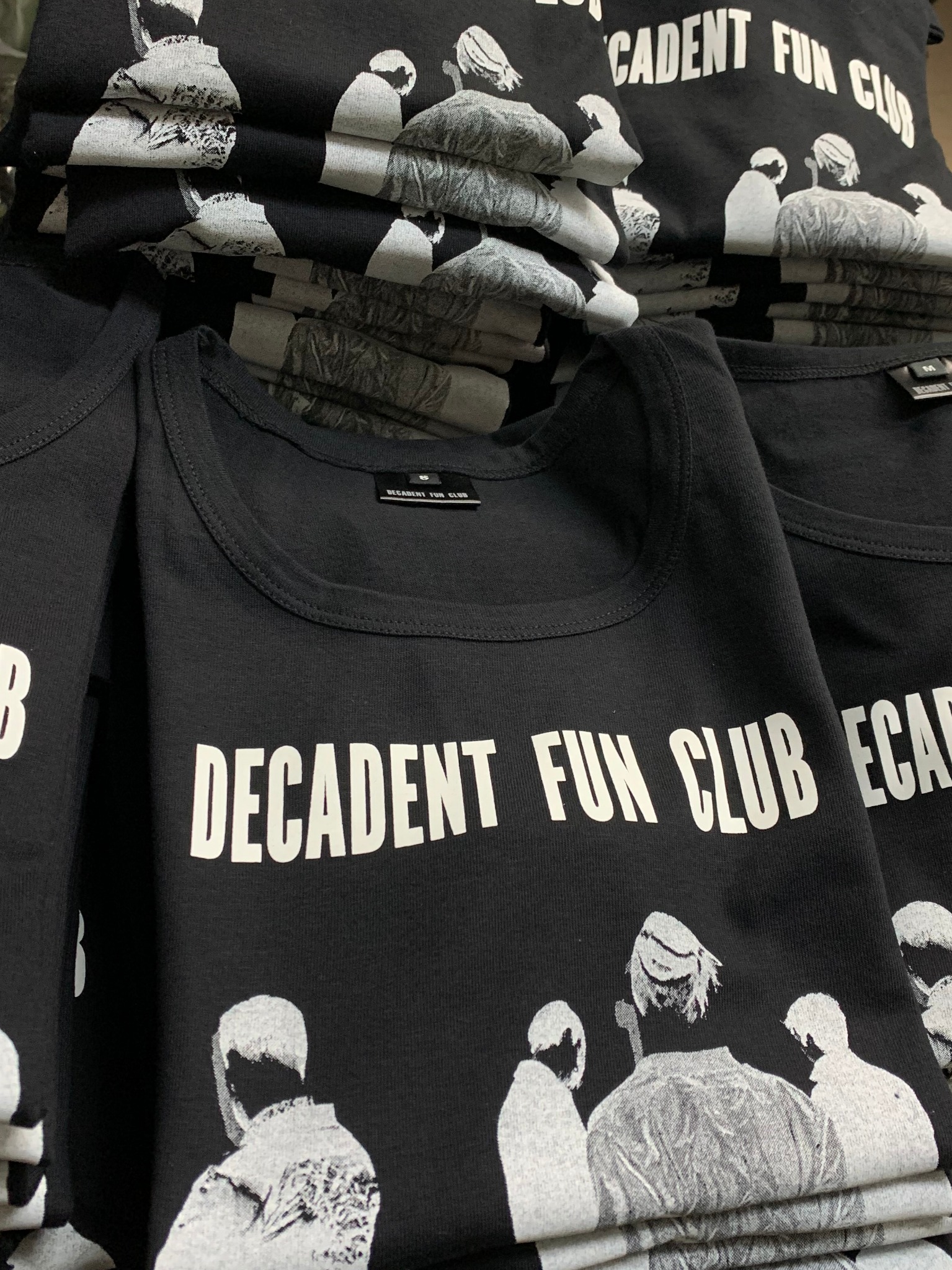 Stos czarnych koszulek z nadrukiem 'Decadent Fun Club', ułożonych w równy stos, z widocznymi detalami nadruku i metkami.