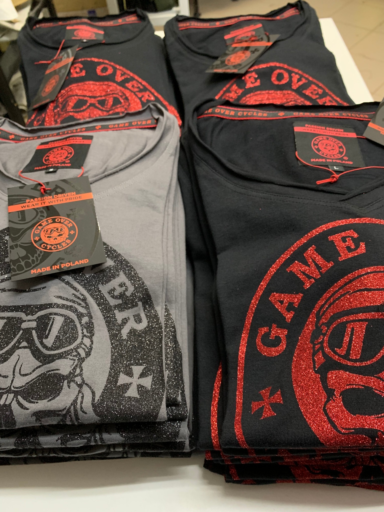 Stos czarnych i szarych koszulek z błyszczącymi nadrukami motocyklowych czaszek i logo 'Game Over Cycles', widoczne metki 'Made in Poland'.