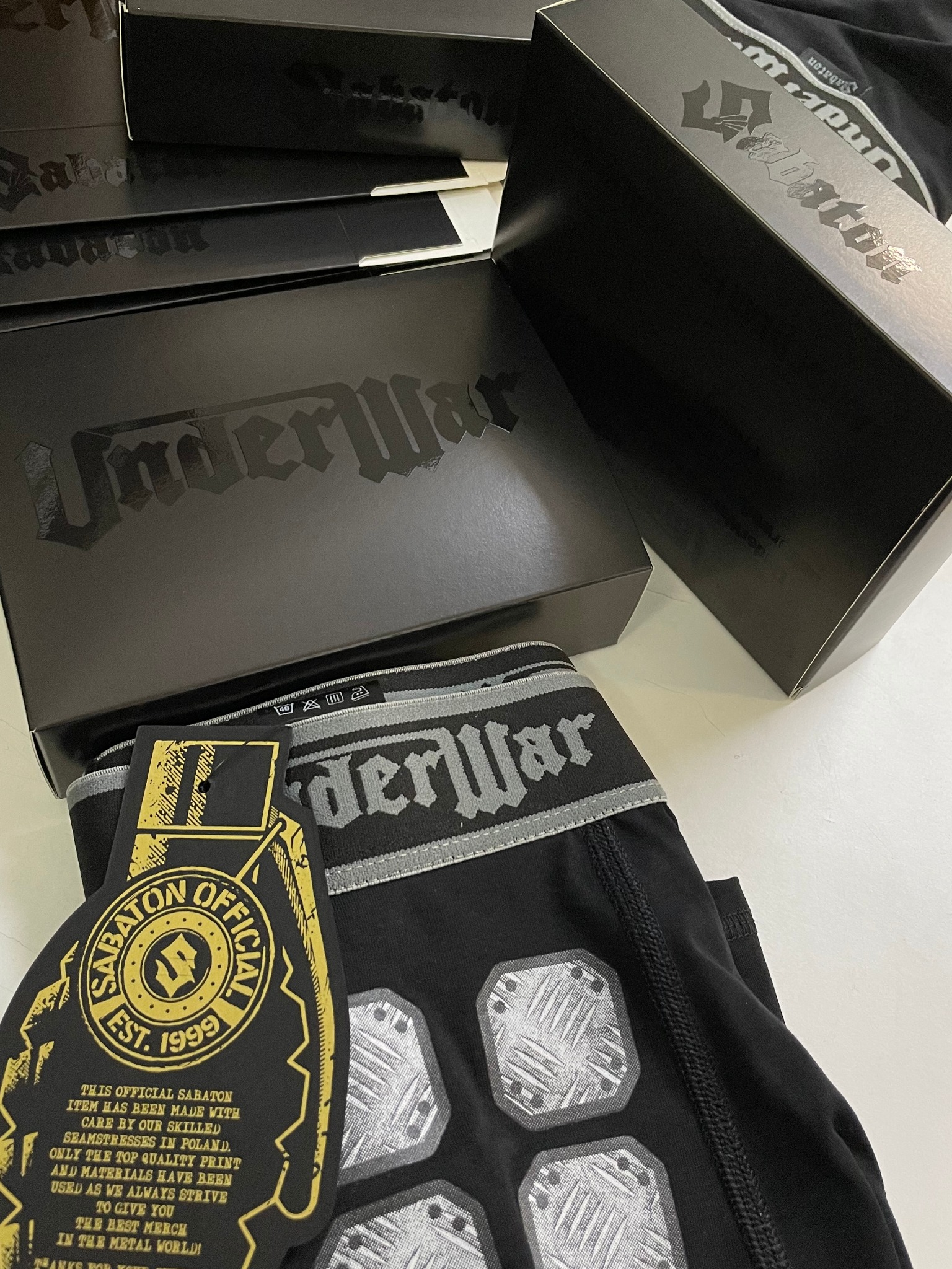 Czarne bokserki UnderWar z szarym pasem i metalicznymi aplikacjami, obok pudełka z logo UnderWar i zawieszką Sabaton Official z informacją o produkcji w Polsce przez wykwalifikowane szwaczki.