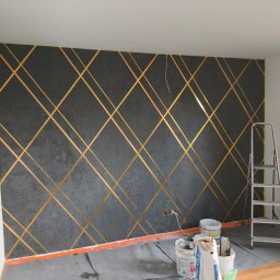 Usługi Remontowo Budowlane Sebastian Korczak - Ściana pomalowana na szaro z geometrycznym wzorem z taśm w kolorze złotym, drabina oparta o ścianę, widoczne puszki z farbą i narzędzia malarskie na podłodze.